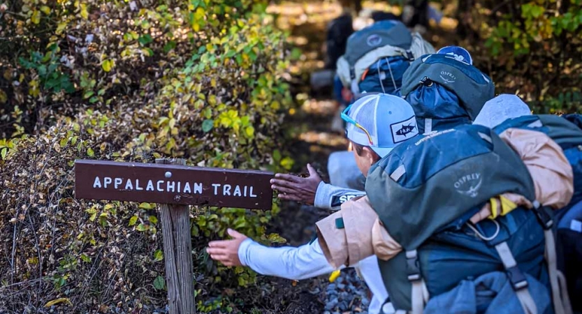 a group of students wearing backpacks pat an Appalachian trail sign on a trip with outward bound
